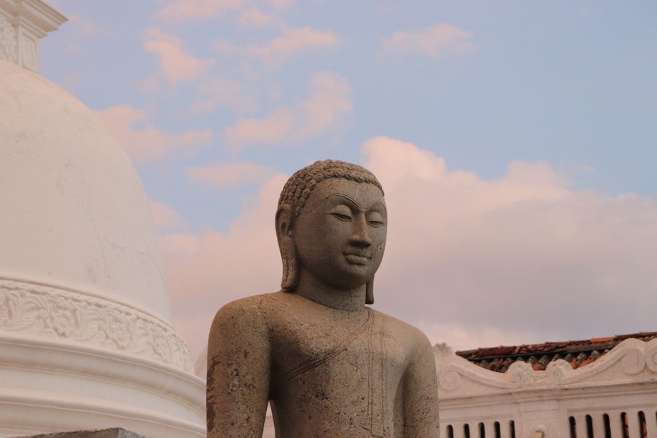A tranquil stone Buddha statue against a vibrant sunrise sky near a white stupa.