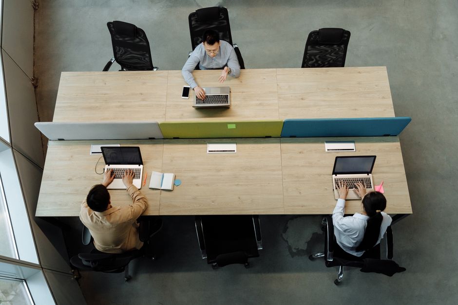 Three colleagues working at a shared office desk, using laptops in a modern workspace.