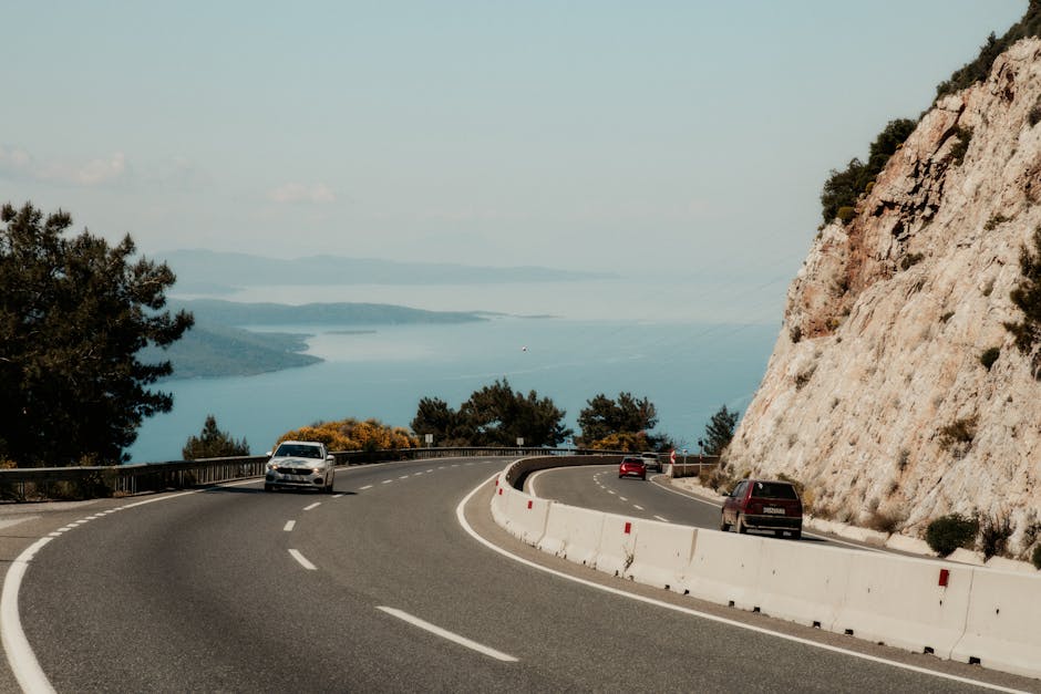 A winding coastal road with cars, overlooking a serene sea and rocky cliffs.
