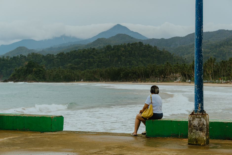A woman sitting by the ocean, enjoying a serene beach with mountains in the background.
