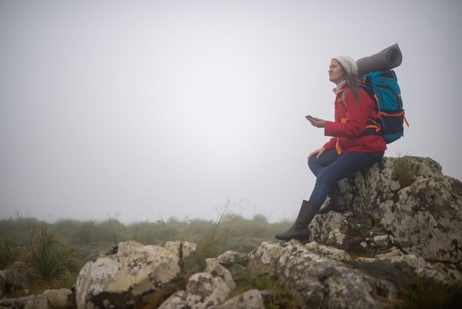 A woman hiker sits on a rock with a backpack and yoga mat in a misty Portuguese landscape.