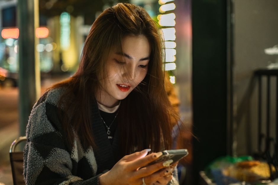 A young woman with brunette hair sits outdoors at night, texting on her phone in an urban setting.