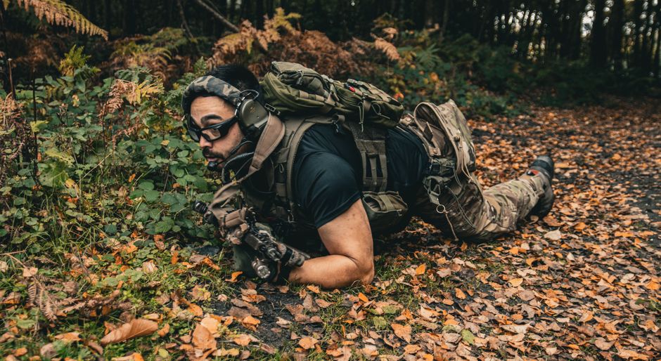 A soldier in full tactical gear crawling through a forest floor covered with autumn leaves.