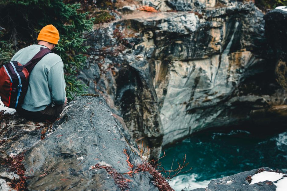 A man with a backpack sits on a rock edge overlooking a scenic canyon in Kootenay Crossing, Canada.
