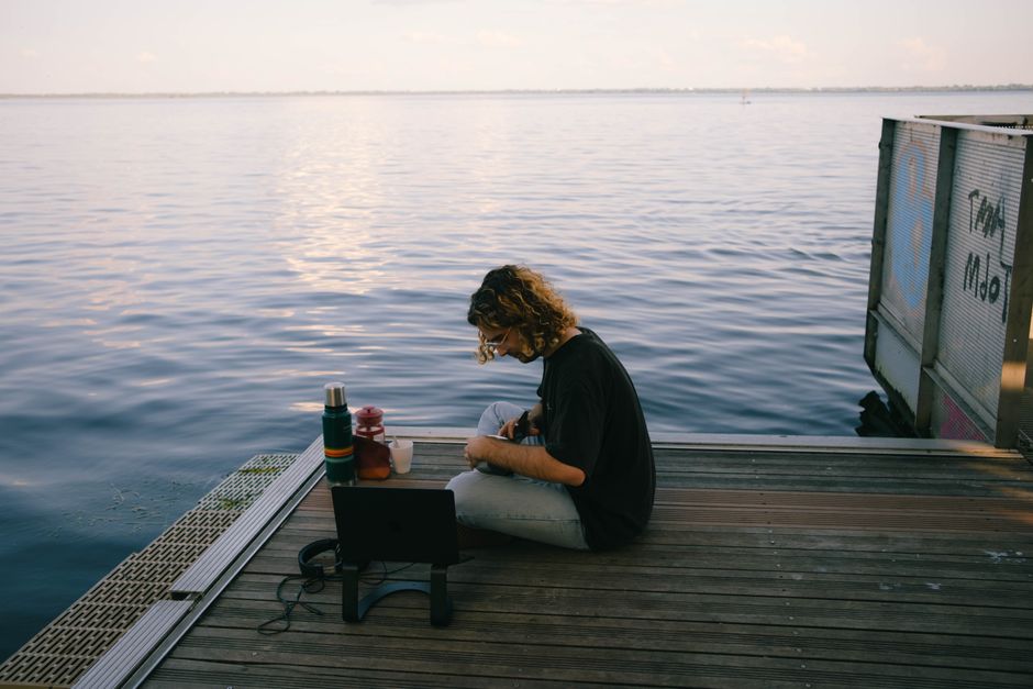 A man sits on a pier, immersed in nature, working on a laptop by the sea.