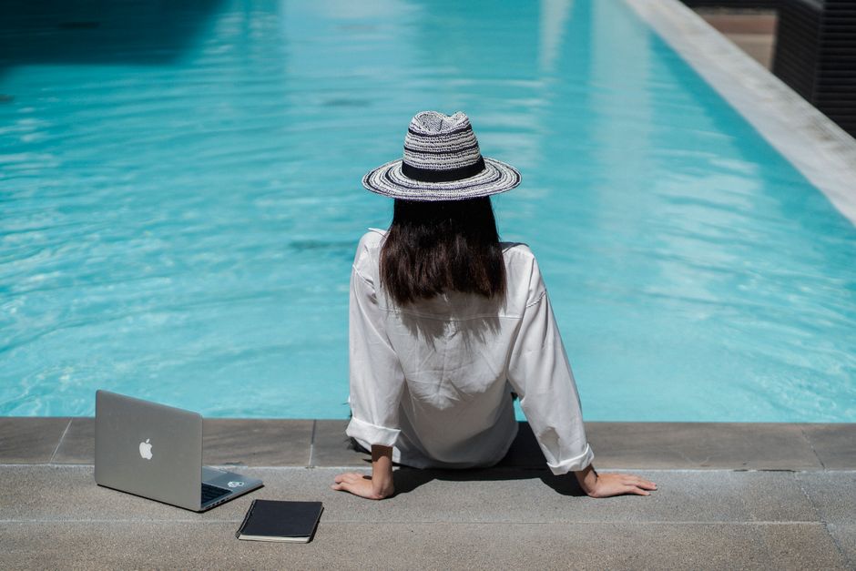 Back view of unrecognizable young self employed woman with dark hair in stylish hat and white shirt relaxing on pool border near opened laptop and notebook