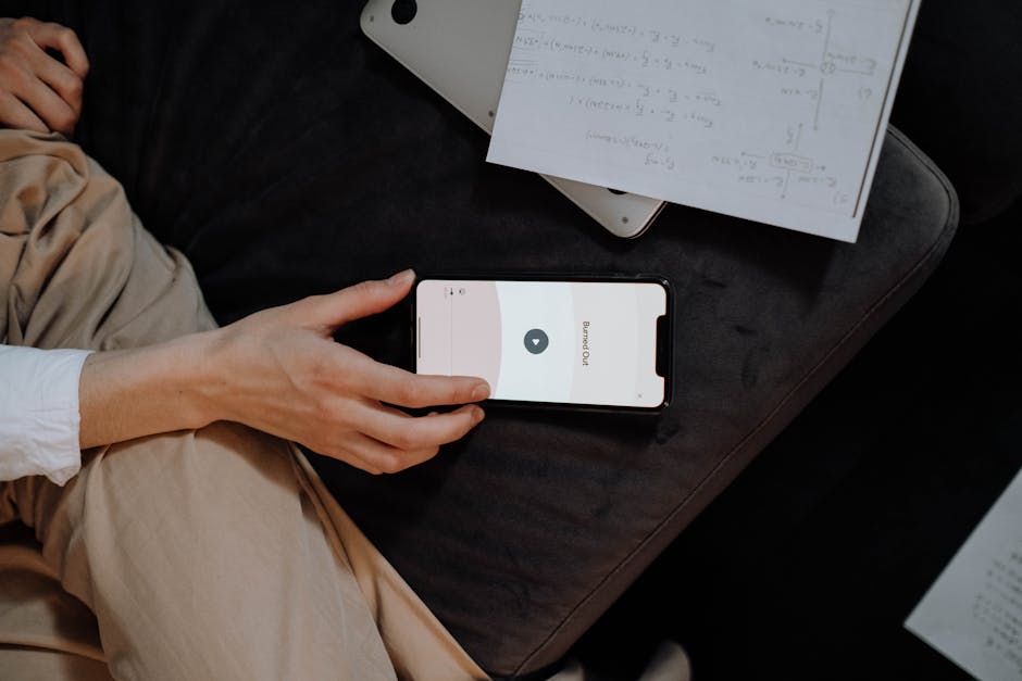 A hand holding a smartphone displaying an app, surrounded by notes and a laptop.