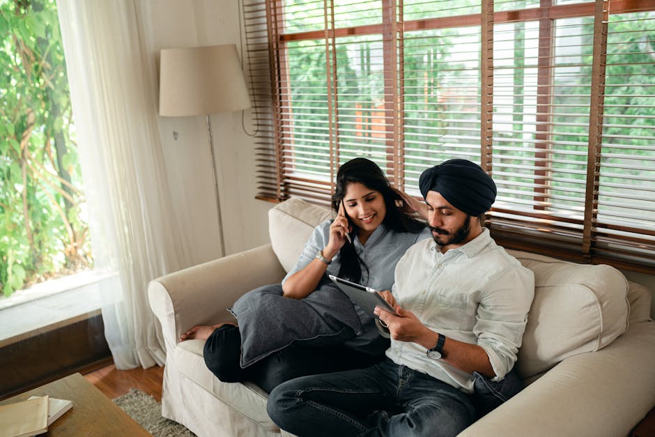 High angle of young Indian woman having phone conversation while husband working on tablet sitting on sofa at home