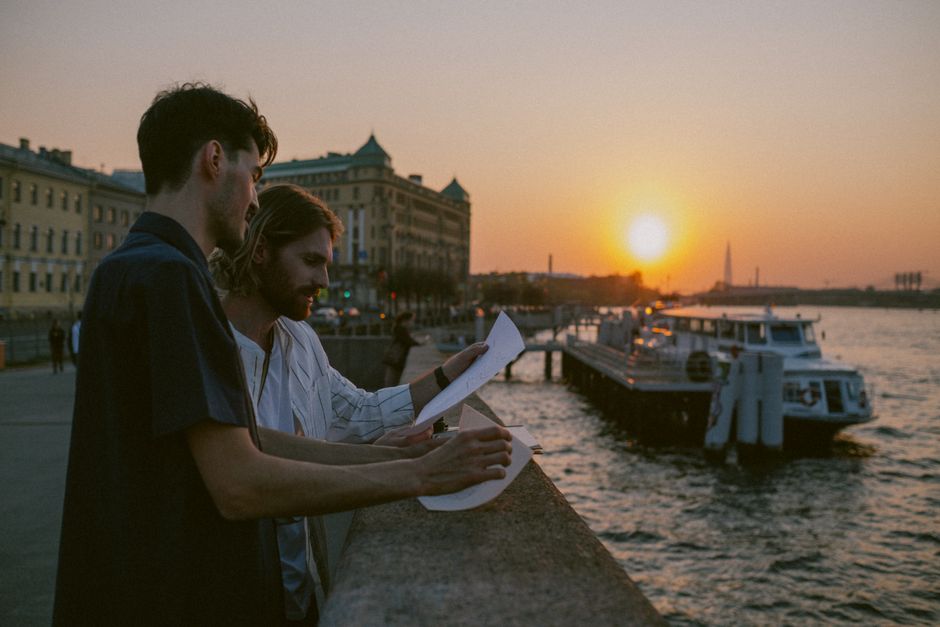 Two men reviewing papers by the riverside with a sunset backdrop, evoking an urban exploration mood.