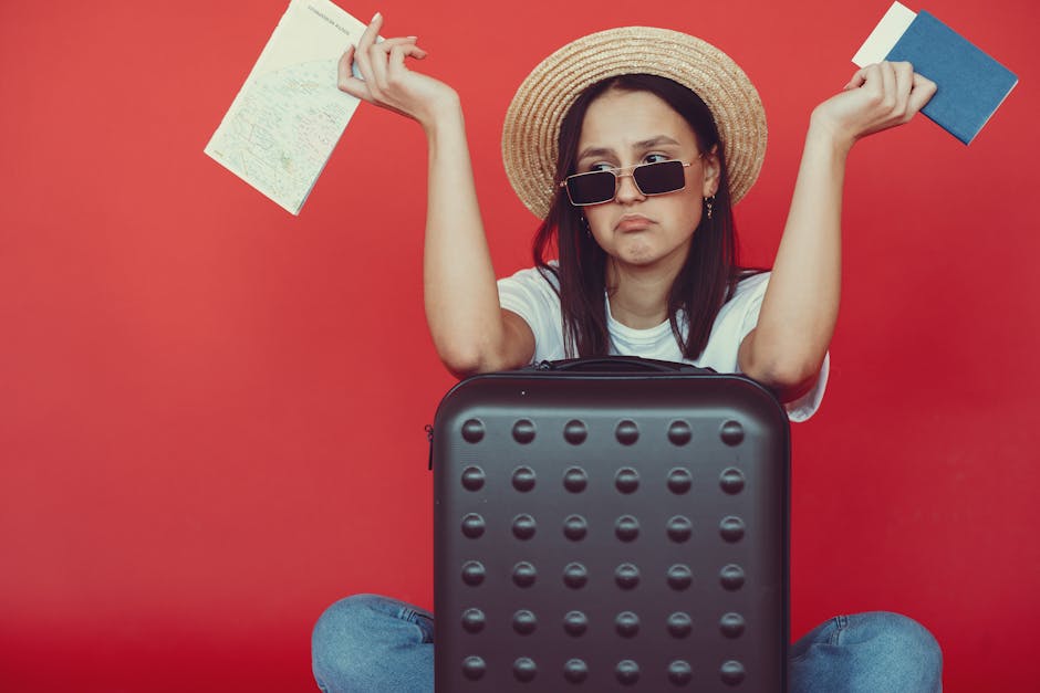 Doubters young stylish woman in wicker hat and trendy sunglasses sitting behind black suitcase and shrugging with passport and tickets on red background