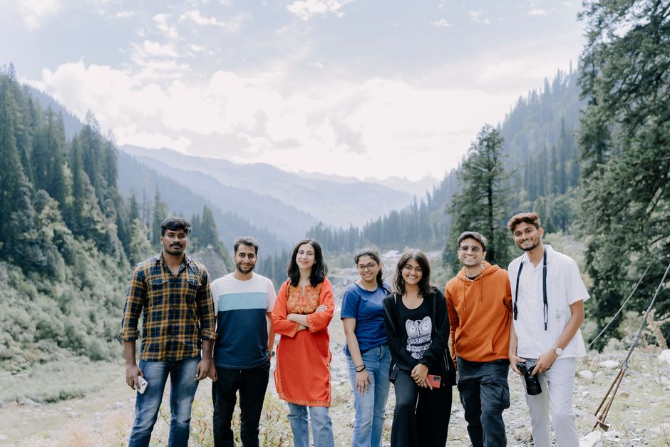 A group of young adults posing together in a scenic mountain landscape, featuring lush greenery and towering trees.
