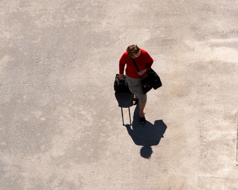 Top-down shot of a woman in red with luggage walking on a paved surface, casting a shadow.