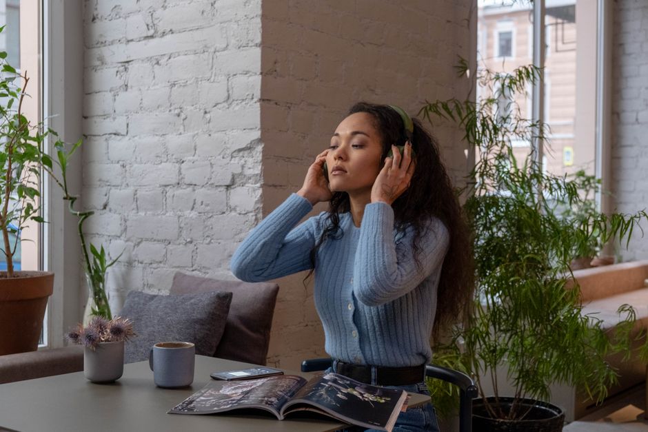 A woman in a blue sweater listens to music with headphones, sitting by a window indoors.