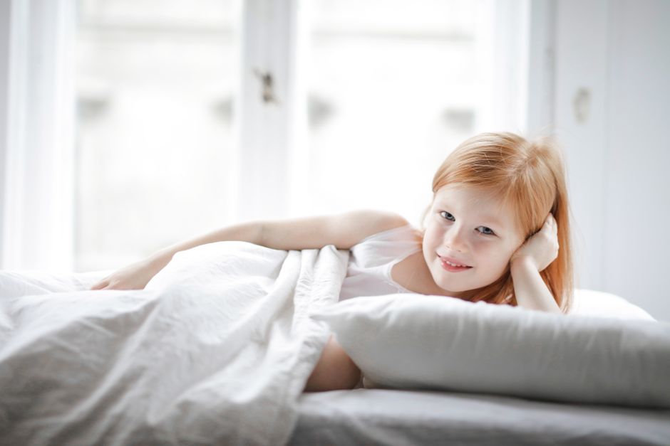 A cheerful young girl with red hair lying on a bed in a bright, cozy bedroom.
