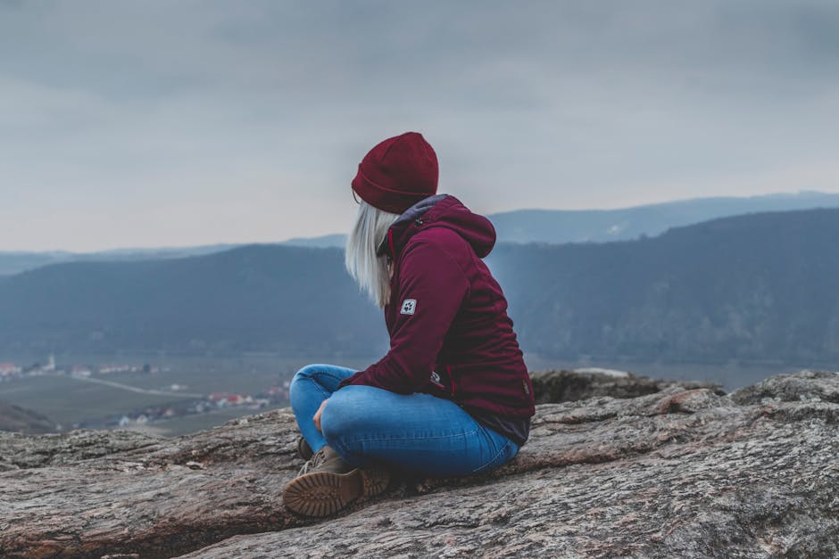 A woman in a maroon jacket sits on a rocky mountain top, gazing at the scenic valley below.